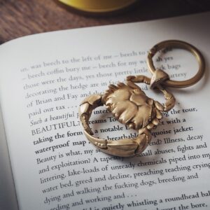 Home Close-up of a golden crab-shaped keychain resting on the pages of an open book, featuring elegant typography.